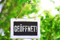 Schild mit der Aufschrift 'Geöffnet!' hängt an einem Baumzweig, umgeben von grünem Laub.