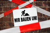 Wir bauen um, steht auf einem Schild vor einer Backsteinmauer und rot-weißem Absperrband eine Baustelle.