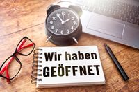 Notizblock mit der Aufschrift 'Wir haben geöffnet' auf einem Schreibtisch, umgeben von einer Brille, einem Wecker und einem Laptop.