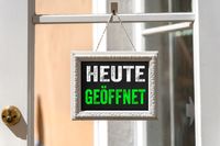 Schild mit der Aufschrift 'Heute geöffnet' in einem weißen Rahmen, das an einer Glastür hängt.