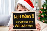 Lustiges Adventsbild: Kind mit Weihnachtsmütze hält eine Tafel mit Aufschrift: Nicht stören! Ich warte auf den Weihnachtsmann.