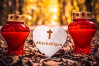 Zwei rote Grabkerzen Herz-Schild mit der Aufschrift „Allerheiligen“ und einem Kreuz-Symbol, auf einem Wald-Friedhof.