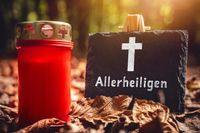 Rote Grabkerze neben einer Tafel mit der Aufschrift „Allerheiligen“ und einem Kreuz-Symbol, umgeben von herbstlichen Blättern.