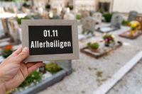 Hand hält ein Schild mit der Aufschrift „01.11 Allerheiligen“ vor einem Friedhof im Hintergrund.