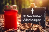 Rote Grabkerze neben einer Tafel mit der Gruß zum „01. November, Allerheiligen“ und einem Kreuz, umgeben von herbstlichen Blättern eines Friedhofs.