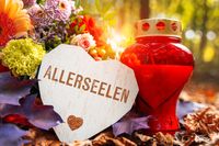 Herzförmiges Schild mit der Aufschrift „Allerseelen“ neben einer roten Grabkerze und bunten Blumengesteck auf einem Grab.