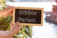 Zwei Hände halten eine Tafel mit der Aufschrift ‚Schöne Ferien!‘ vor einer sonnigen Landschaft, passend zu den Faschingsferien 2025.