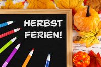 Tafel mit der Aufschrift ‚Herbstferien!‘, umgeben von bunten Herbstblättern und Kürbissen, passend zu den Herbstferien 2025.