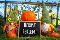 Zwei Stoffzwerge mit Kürbissen und einer Tafel mit der Aufschrift ‚Herbstferien‘ im Garten, passend zu den Herbstferien 2025.