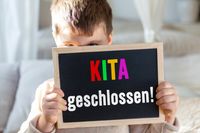 Ein Kind hält eine Tafel mit der Aufschrift ‚KITA geschlossen‘, passend zur Situation, wenn der Kindergarten geschlossen ist.