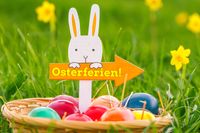 Schild in Form eines Osterhasen mit der Schriftzug ‚Osterferien‘ neben bunt bemalten Ostereiern im Gras, passend zu den Osterferien 2025.