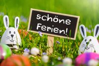 Tafel mit der Aufschrift ‚schöne Ferien!‘ im Gras, umgeben von Osterhasen und bunten Eiern, passend zu den Osterferien 2025 an Ostern.