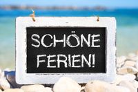 Tafel mit der Aufschrift ‚Schöne Ferien!‘ auf einem Kiesstrand mit Meer im Hintergrund, passend zu den Pfingstferien 2025.