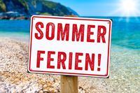 Schild mit der Aufschrift ‚Sommerferien!‘ am Strand mit Meer im Hintergrund, passend zu schönen Sommerferien 2025.