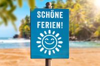 Blaues Schild mit der Feriengruß ‚Schöne Ferien!‘ und einem lachenden Sonnensymbol am Strand, das den Beginn schöner Ferien signalisiert.