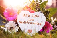 Alles Liebe zum Weltfrauentag! steht auf einem herzförmigen Schild, das in einem Blumenarrangement mit rosa und weißen Gerbera liegt.