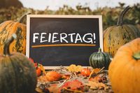 Feiertag! steht auf einem kleinen Schild, das zwischen Kürbissen und Herbstlaub im Freien aufgestellt ist.