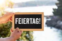 Feiertag! steht auf einem kleinen Schild, das von einer Person vor einer sonnigen Landschaft mit Bäumen und Wasser im Hintergrund gehalten wird.