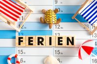 Ferien! steht auf Holzbuchstaben, die auf einem Kalender liegen, umgeben von Strandmotiven wie Liegestühlen, einer Schildkröte und einem Rettungsring.