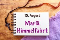 Buch mit dem Text ‚15. August Mariä Himmelfahrt‘, daneben ein schwarzer Rosenkranz auf einem Holztisch. Das Bild erinnert an den Feiertag im Sommer.