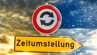 Verkehrsschild mit Pfeilen und einem gelben Schild mit der Aufschrift ‚Zeitumstellung‘. Symbol für das Umstellen der Uhren.