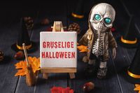 Gruselige Bilder Halloween – Detailaufnahme eines Skeletts mit leuchtenden Augen, umgeben von Halloween-Dekoration, wie kleinen Hexenhüten und Besen, auf einem dunklen Hintergrund.