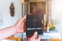 Hände halten ein schwarzes Buch mit der Aufschrift „Mariä Himmelfahrt“ und einem Kreuz, im Hintergrund ist ein unscharfer Altar in einer Kirche zu sehen.