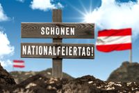 Schild mit der Aufschrift „Schönen Nationalfeiertag!“ In Österreich, vor einem blauen Himmel mit Wolken, im Hintergrund zwei unscharfe österreichische Fahnen zu sehen.