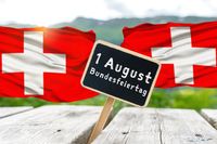 Zwei Schweizer Fahnen wehen, im Vordergrund ein Schild mit der Aufschrift „1. August Bundesfeiertag“ auf einem Holztisch steht, symbolisiert Nationalfeiertag der Schweiz.