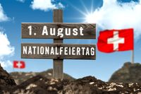 Holzschild mit der Aufschrift „1. August Nationalfeiertag“ Schweiz Fahnen vor einem blauen Himmel.