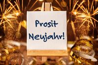 Prosit Neujahr. Eine Staffelei mit der Aufschrift Prosit Neujahr, im Hintergrund Feuerwerk und Deko.