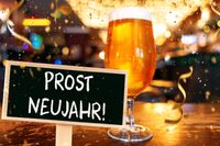 Prost Neujahr. Ein Glas Bier steht auf einem Tisch, daneben ein Schild mit der Aufschrift Prost Neujahr.