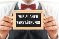 Wir suchen Verstärkung! Steht auf einem Schild, dieses wird von einem Kellner in Uniform gehalten – Symbol für Jobangebote.