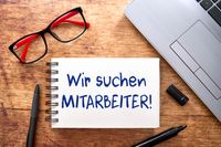 Notizblock mit der Aufschrift 'Wir suchen Mitarbeiter!' auf einem Schreibtisch mit Brille und Laptop, Mitarbeiter gesucht.