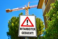 Ein Schild mit der Aufschrift „Mitarbeiter gesucht!“ steht vor einer Baustelle mit einem Kran im Hintergrund, was auf eine Stellenausschreibung im Handwerk oder für Techniker hinweist.