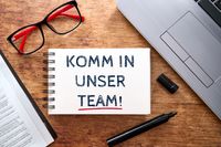 Komm in unser Team, steht auf einem Buch, umgeben von einem Laptop, einer Brille und einem Stift im Büro. Themenbild zu Stellenausschreibung im Bereich Büromanagement hin.