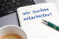 Ein Notizblock mit der Aufschrift „Wir suchen Mitarbeiter!“ liegt auf einem Schreibtisch neben einem Laptop und einer Tasse Kaffee, symbolisch für eine Stellenausschreibung für Bürokräfte.