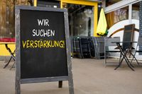 Tafel vor einem Restaurant mit der Aufschrift „Wir suchen Verstärkung!“ Bild für Suche nach Mitarbeitern über Stellenanzeige im Gastronomiebereich, z.B. Koch, Kellner, Servicekraft.