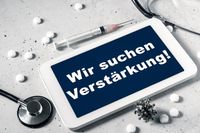 Ein Tablet mit der Aufschrift „Wir suchen Verstärkung!“ liegt auf einem Tisch, umgeben von einem Stethoskop, einer Spritze und Tabletten. Dies deutet auf eine Stellenanzeige im medizinischen Bereich hin, insbesondere für Alten- und Krankenpfleger oder medizinische Fachangestellte.