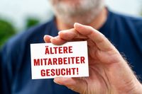 Ältere Mitarbeiter gesucht! Schild in einer Hand, welches auf eine Stellenausschreibung für erfahrene Arbeitskräfte und Senioren hinweist.