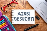 Azubi gesucht! Steht auf einem Schulheft, welches auf einem Schreibtisch liegt, umgeben von Stiften, einer Brille und einem Laptop, was auf eine Stellenausschreibung für Azubis hinweist.