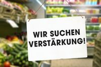 Ein Schild mit der Aufschrift „Wir suchen Verstärkung!“ hängt vor einem Verkaufsbereich, was auf eine Stellenausschreibung im Handel hinweist.