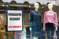 Ein Schild mit der Aufschrift „Verkäufer gesucht, jetzt bewerben!“ weist auf eine Stellenanzeige im Einzelhandel hin. Schaufenster mit zwei Schaufensterpuppen in Kleidung.