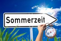 Ein Straßenschild mit der Aufschrift 'Sommerzeit' und eine Hand, die einen Wecker hält – Symbol für das Uhren umstellen zur Sommerzeit.