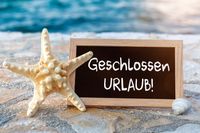 Ein kleines Schild mit der Aufschrift „Geschlossen - Urlaub!“ steht auf einem Steinboden neben einem Seestern und einer Muschel, mit dem Meer im Hintergrund.