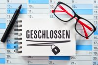 Geschlossen, steht auf einem Notizbuch, der auf einem Kalender liegt, daneben eine Brille und ein Stift.