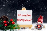 Schild, auf dem „Wir machen Winterpause!“ steht, umgeben von winterlicher Dekoration mit Schneeflocken, Tannenzweigen und einem kleinen gestrickten Figurenornament auf Skiern.