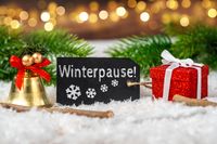 Winterpause auf einer Tafel, umgeben von weihnachtlicher Dekoration, Geschenken und Tannenzweigen im Schnee, symbolisch für den Winterurlaub.