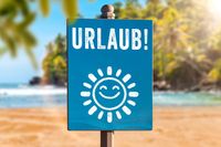 Ein blaues Schild mit der Prägung „Urlaub!“ und einem lächelnden Sonnen-Symbol, das auf Sommerpause und Sommerurlaub hinweist, vor Strandkulisse.