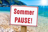 Sommerpause aufgeschrieben auf einem Schild zum Urlaub am Strand mit blauem Meer im Hintergrund, symbolisch für Sommerurlaub.